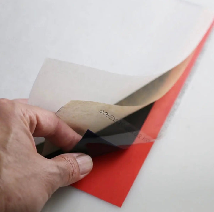 Hand peeling back the layers of Carbon &amp; Ribbon stencil paper, revealing the carbon transfer sheet and red backing paper.